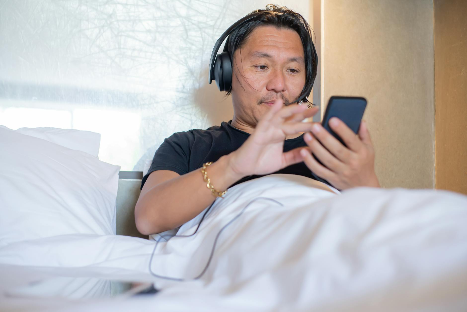 Azjata słuchający audio w słuchawkach i telefonie w przytulnej sypialni